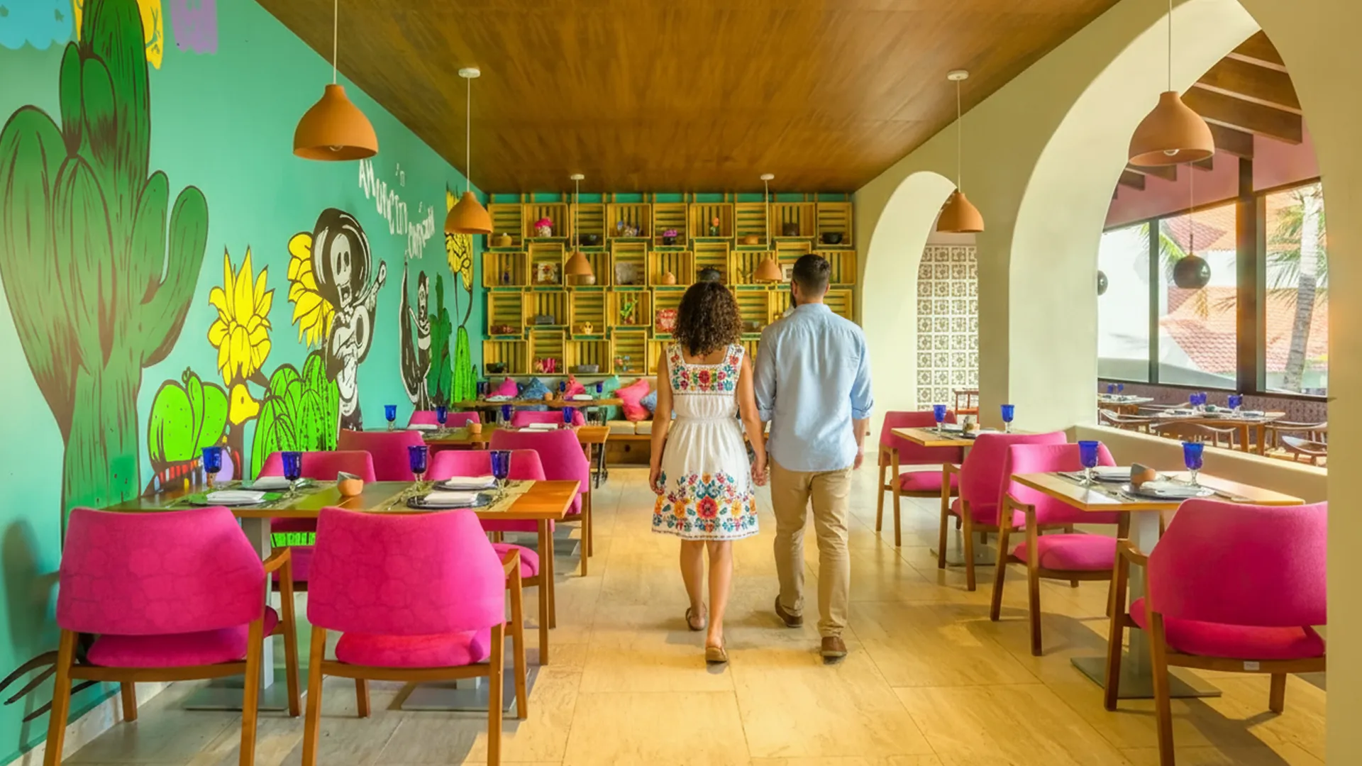A couple walking through a colorful Mexican-themed restaurant in a Cancun resort, featuring vibrant turquoise walls with cartoonish cactus, skull, and monkey murals, wooden shelves stocked with pottery, pink upholstered chairs at wooden tables set with blue glasses, terracotta pendant lamps, and arched doorways leading to an outdoor patio with sunset views.