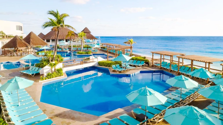Aerial view of a tropical resort featuring a large circular pool surrounded by palm trees, thatched-roof structures, lounge chairs, and a beachfront with the ocean in the background.