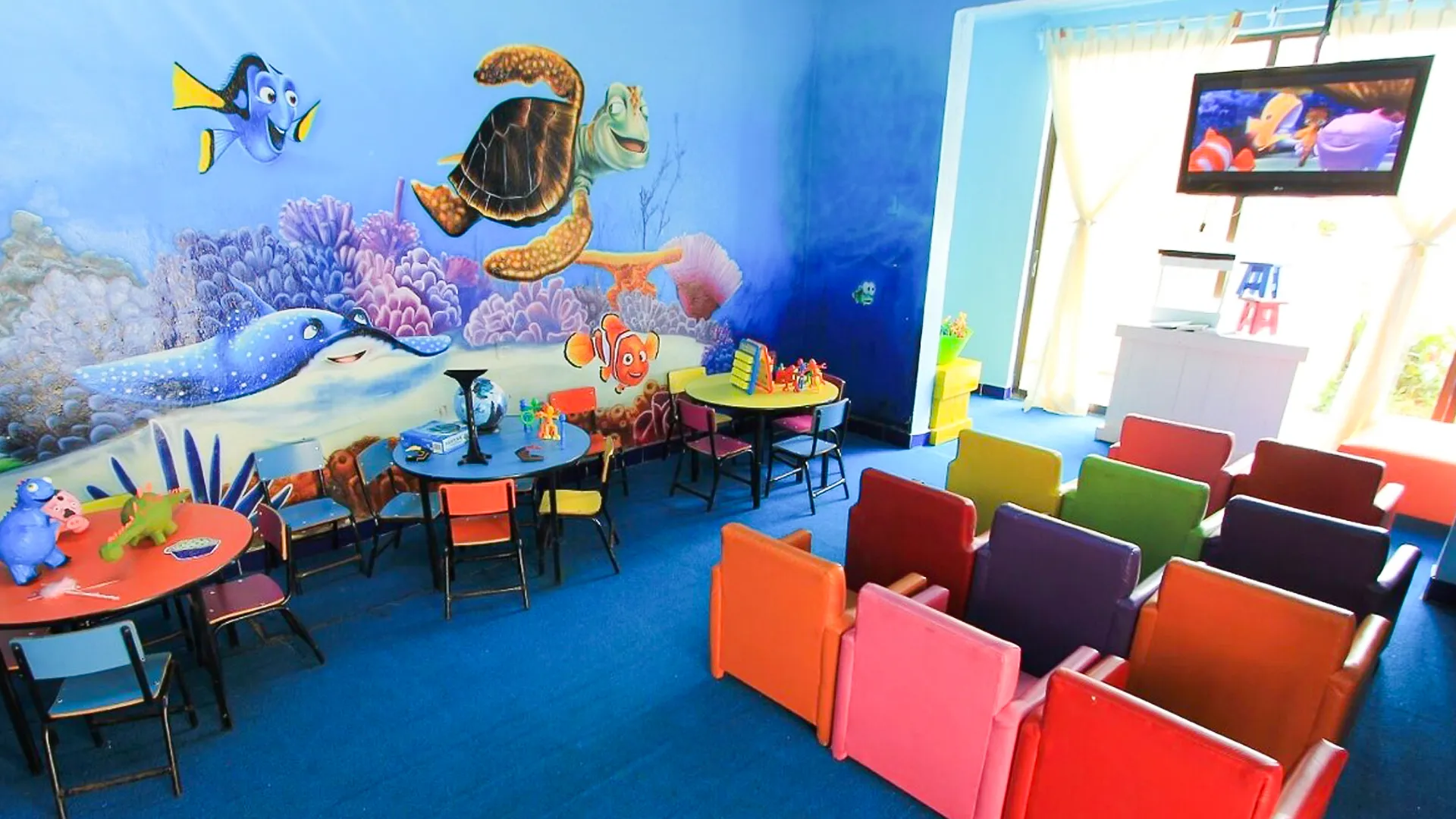 Colorful indoor kids’ club at All Ritmo Cancun Resort featuring ocean-themed wall art, mini chairs, and a movie screen showing an animated film.