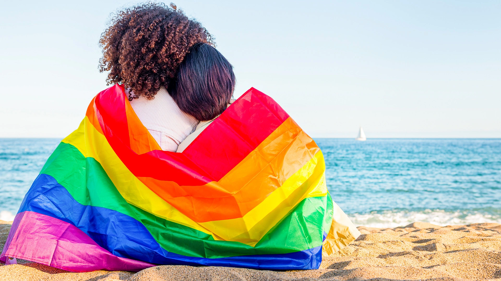 Two people wrapped in a rainbow pride flag embracing on the beach with the ocean in the background.