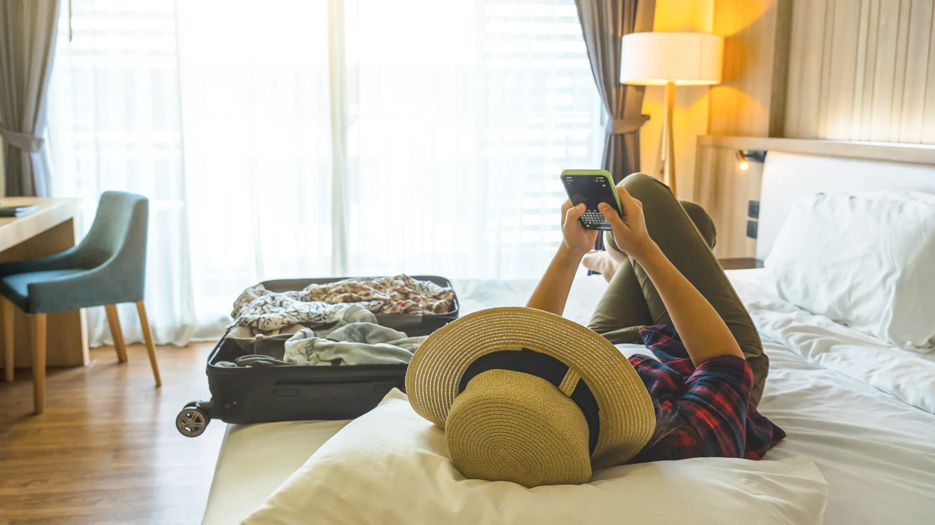 Woman lying on a hotel bed with a straw hat and plaid shirt, legs up, browsing a green smartphone while packing clothes spill from an open black suitcase nearby, soft room lighting.