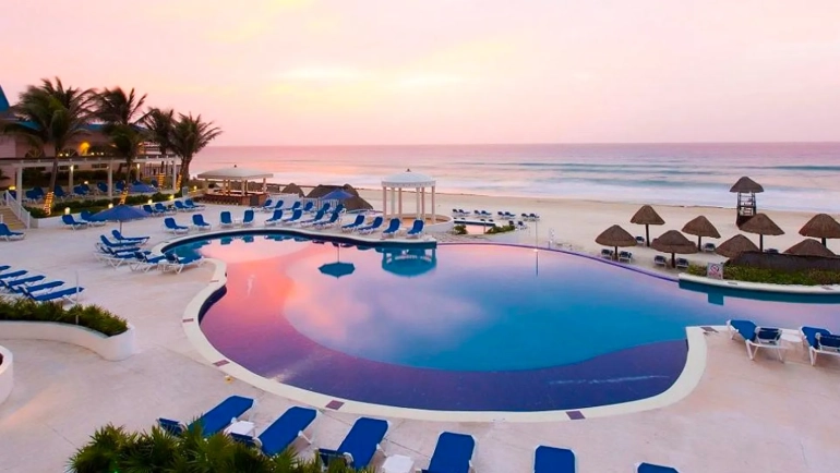 A view from a balcony overlooking a sandy beach with blue lounge chairs, thatched-roof umbrellas, and the turquoise ocean, with tables and chairs on the balcony in the foreground.