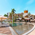 A vibrant pool area at Villa del Palmar Cancun Resort featuring a large infinity pool, white lounge chairs, beige umbrellas, palm trees, and a colorful multi-story building under a clear blue sky.