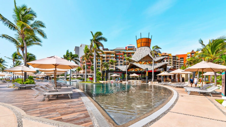 A vibrant pool area at Villa del Palmar Cancun Resort featuring a large infinity pool, white lounge chairs, beige umbrellas, palm trees, and a colorful multi-story building under a clear blue sky.