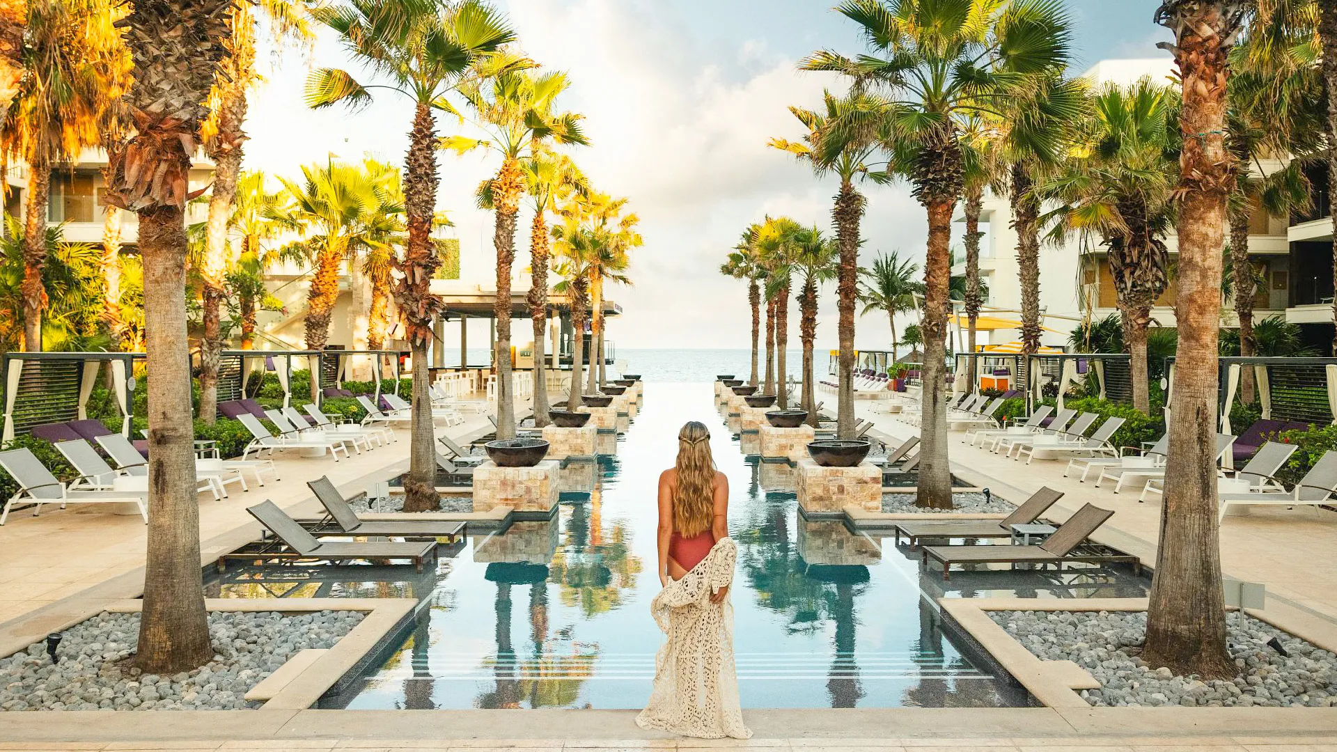 Woman standing by the palm-lined infinity pool at Breathless Riviera Cancun Resort during sunset.
