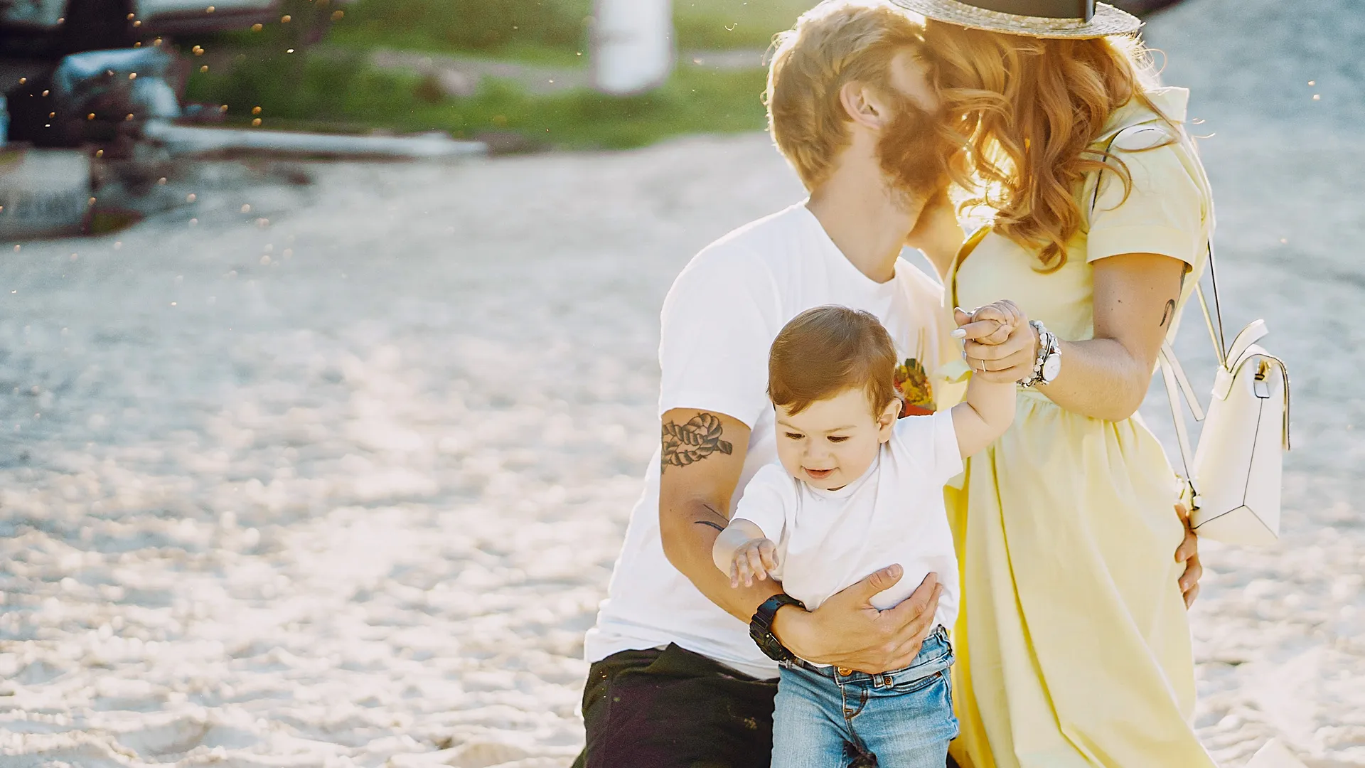 Heartwarming family portrait on the beach at Crown Paradise Club Cancun, with a father in a white shirt and jeans kneeling to hug his wife in a yellow sundress and their young son in a white outfit and jeans, all smiling under golden hour light with white sand and gentle waves behind them.