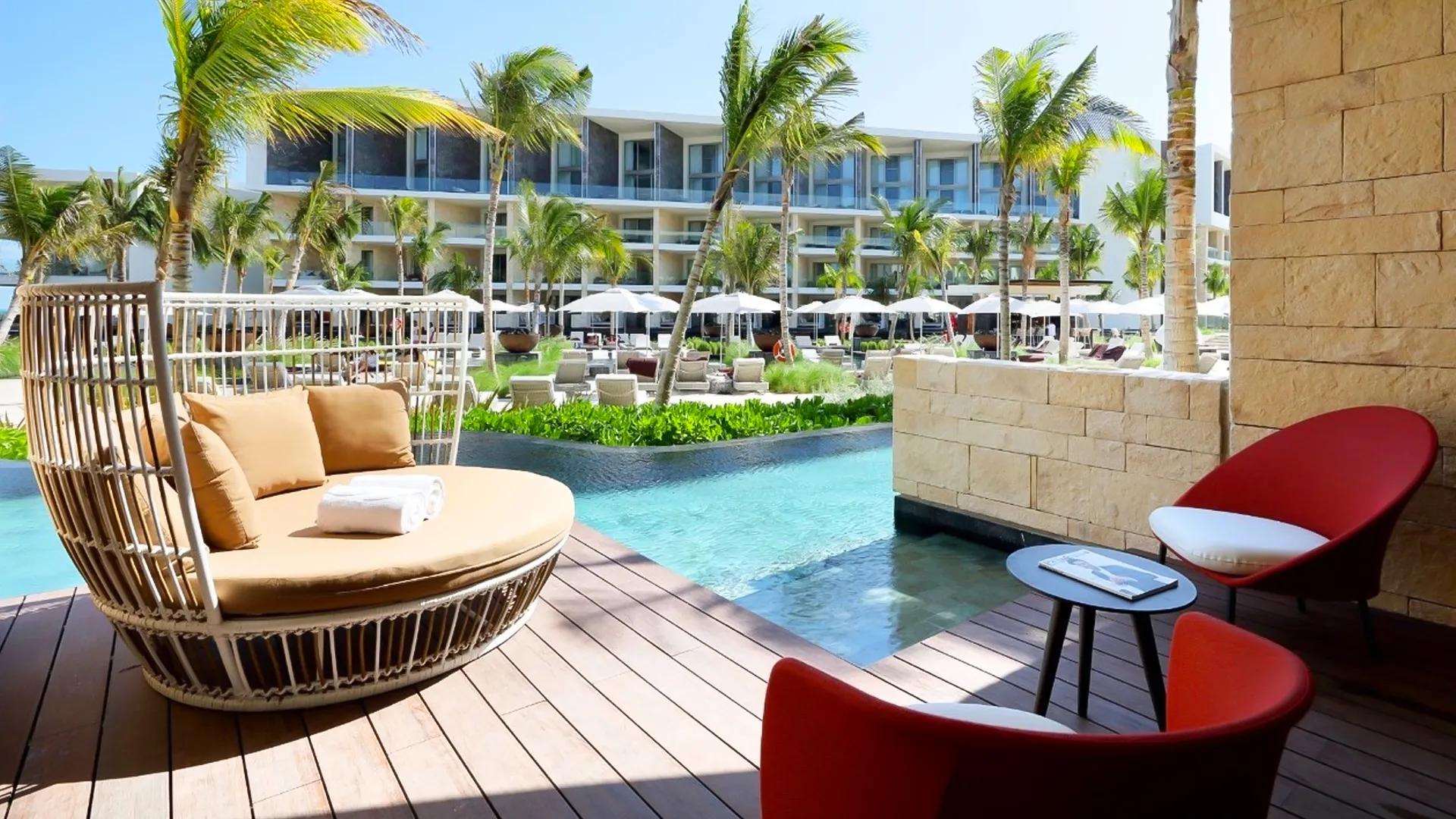 A private patio at Grand Palladium Cancun Resort with a wooden deck, a cushioned wicker chair, a red modern chair, a small table, and a view of the pool and resort buildings with palm trees in the background.