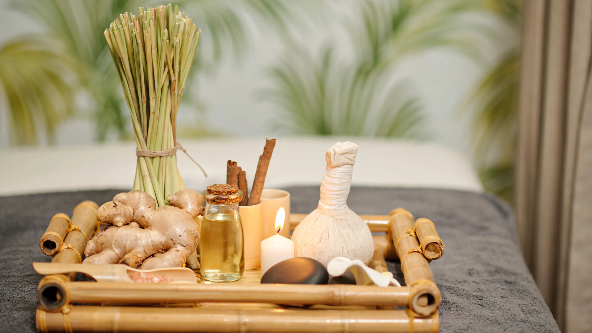 A spa tray at Grand Palladium Cancun Resort containing ginger roots, a bottle of oil, cinnamon sticks, a lit candle, a hot stone, and a muslin pouch, set against a backdrop of green palm wallpaper and a massage table.