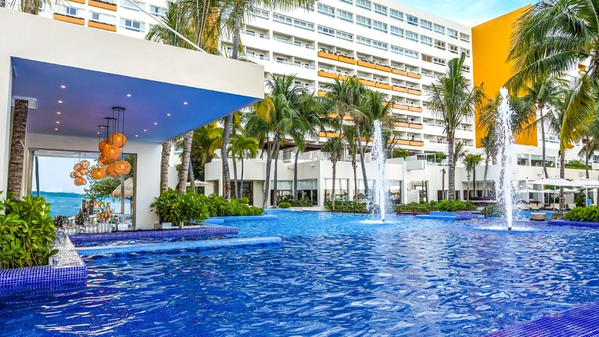 A vibrant multi-level pool area at a Cancun resort, with cascading blue-tiled infinity pools, illuminated fountains spraying water, lush green plants, orange lanterns hanging from a white pergola, tall palm trees, and white multi-story resort buildings in the background under a partly cloudy sky.