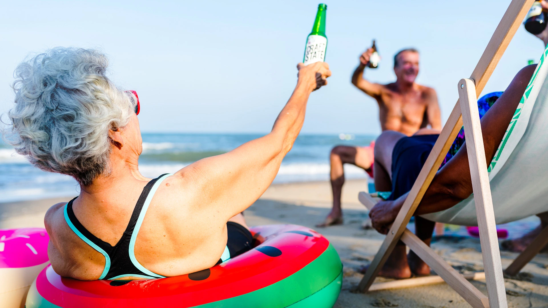 Older couple enjoying beers and laughter on the beach, relaxing on sun chairs by the sea.
