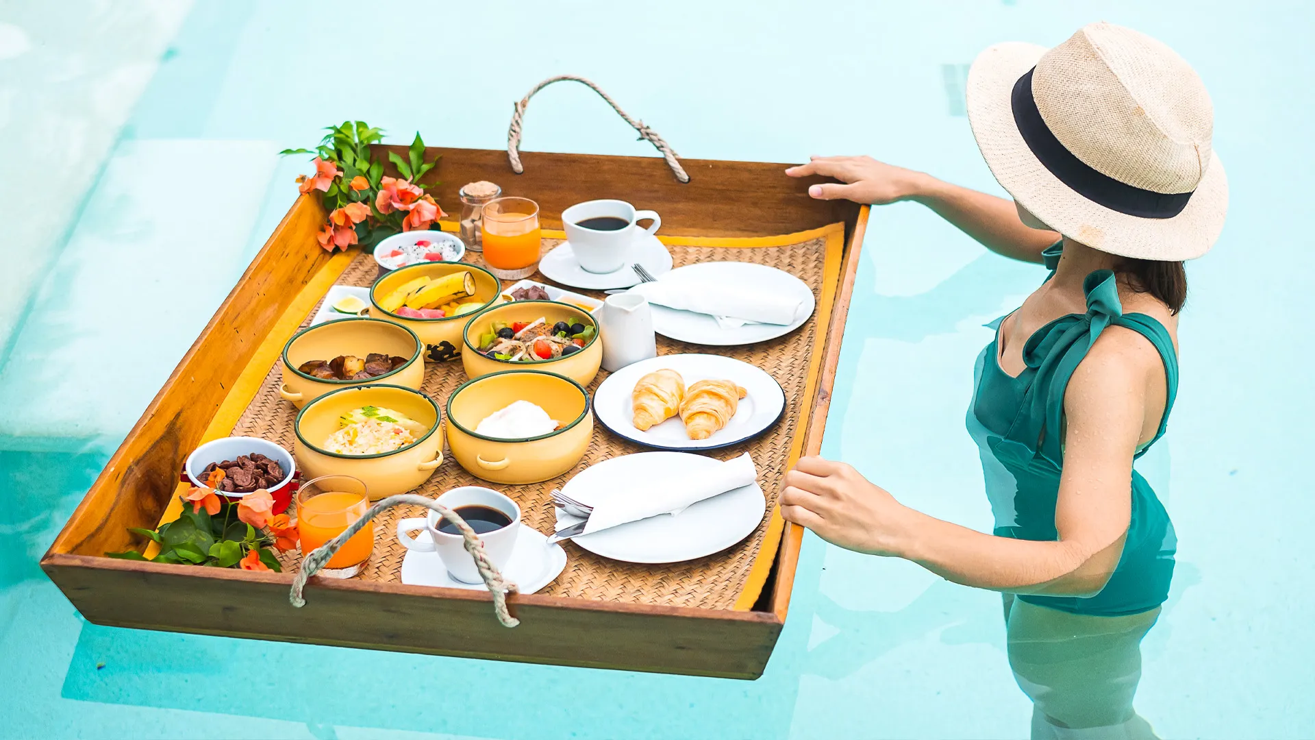 Woman in a straw hat enjoying a floating breakfast tray with tropical fruits, pastries, and coffee in a resort pool.