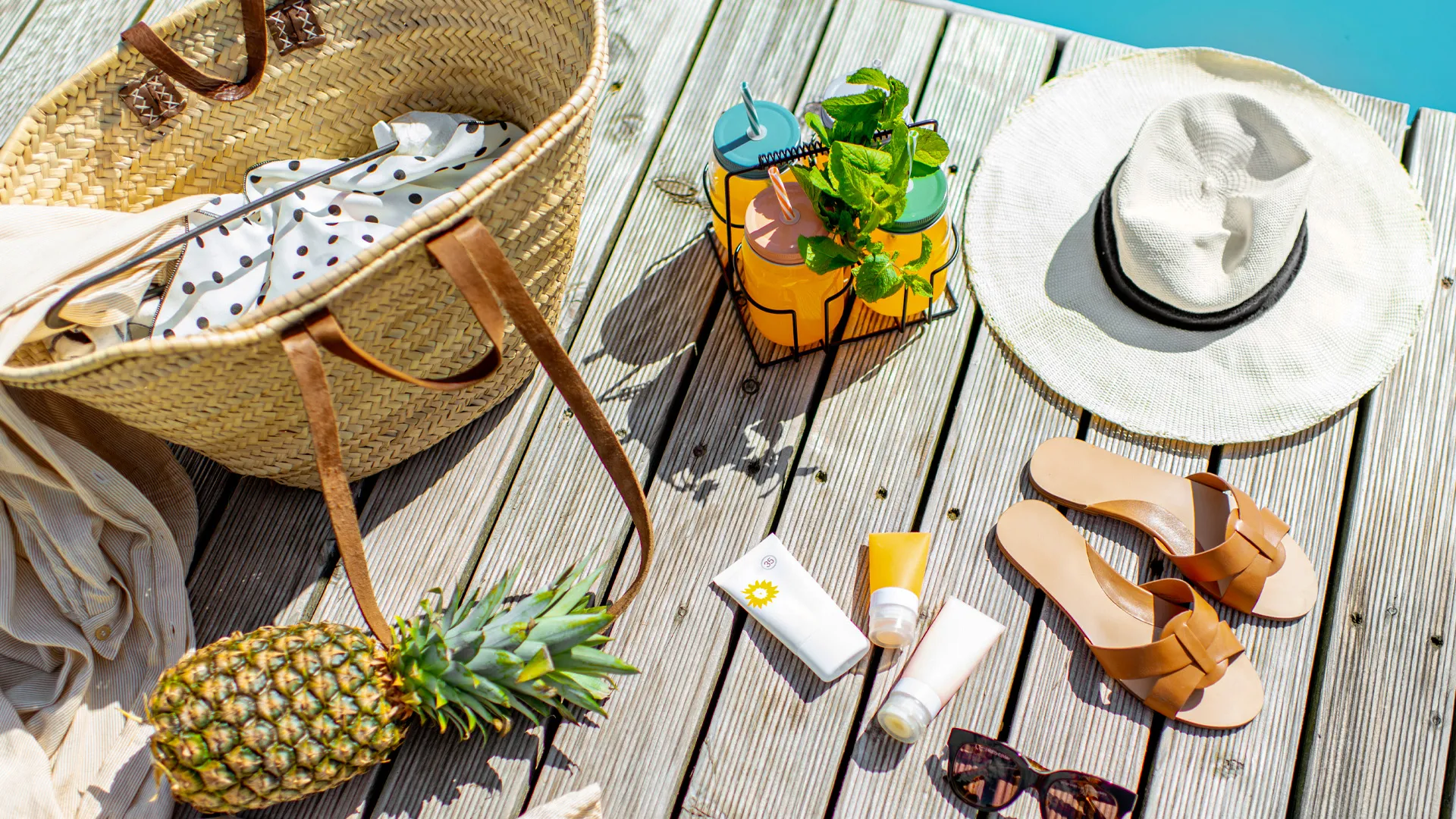 Summer flat lay on a wooden deck with a straw beach bag, pineapple, sun hat, sandals, sunglasses, and tropical drinks by the pool.