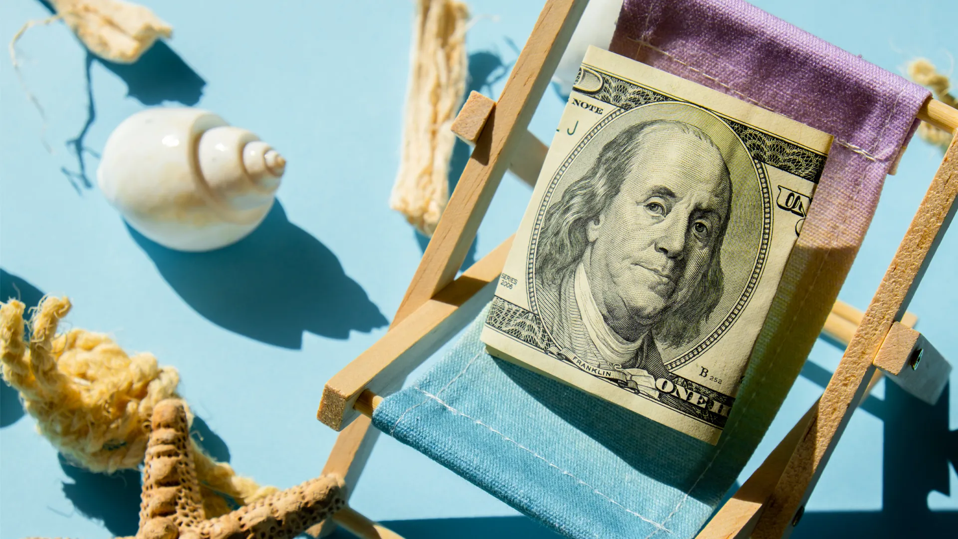 Rolled-up US dollar bill placed on a miniature beach chair with seashells and coral decoration.