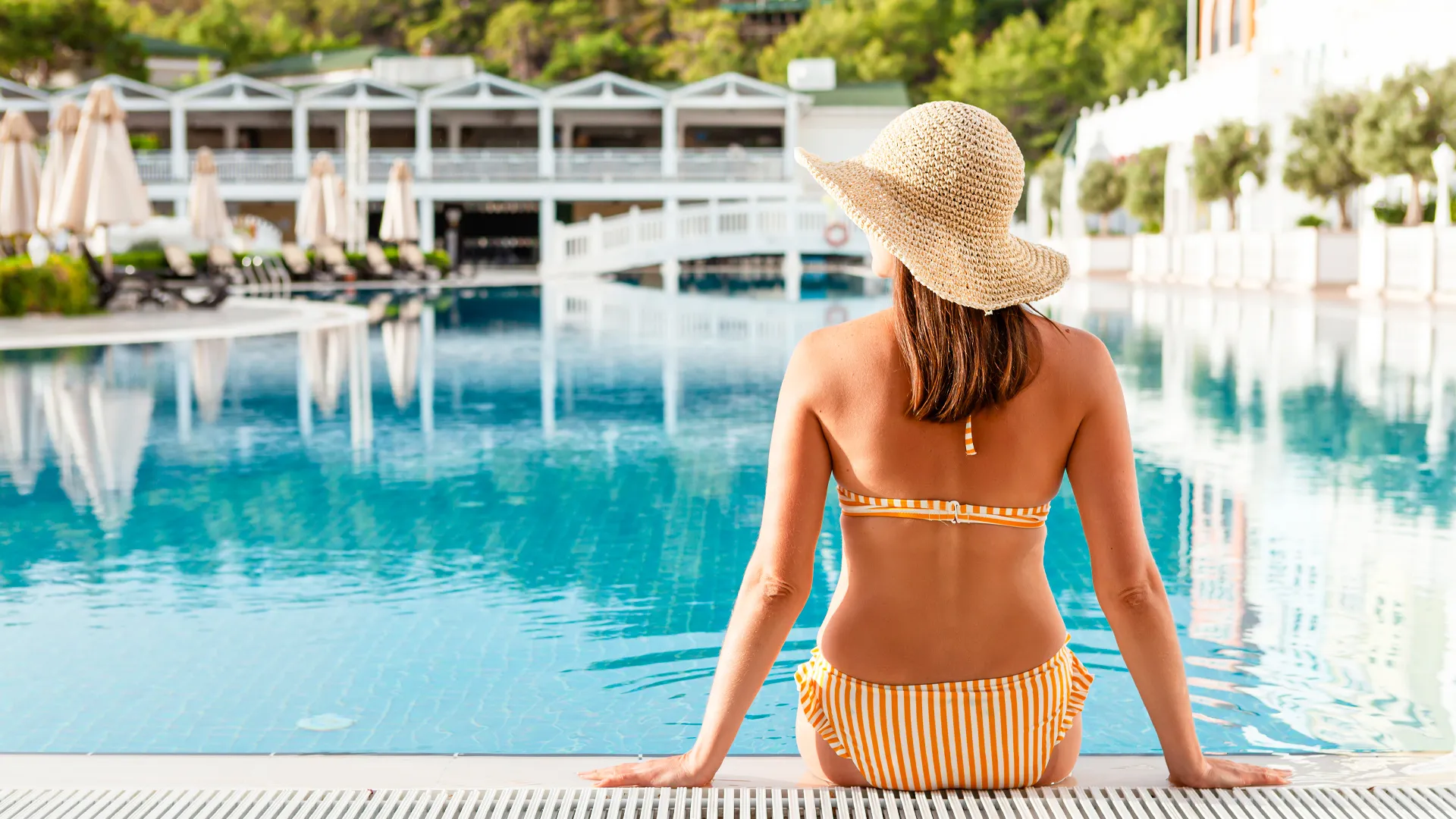 Woman in striped bikini and straw hat sitting by a luxury resort pool surrounded by sunbeds and umbrellas.