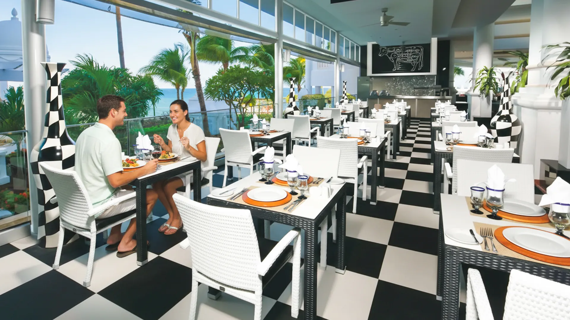 Couple dining at the elegant ocean-view restaurant at Riu Palace Cancun with black-and-white décor and palm views.