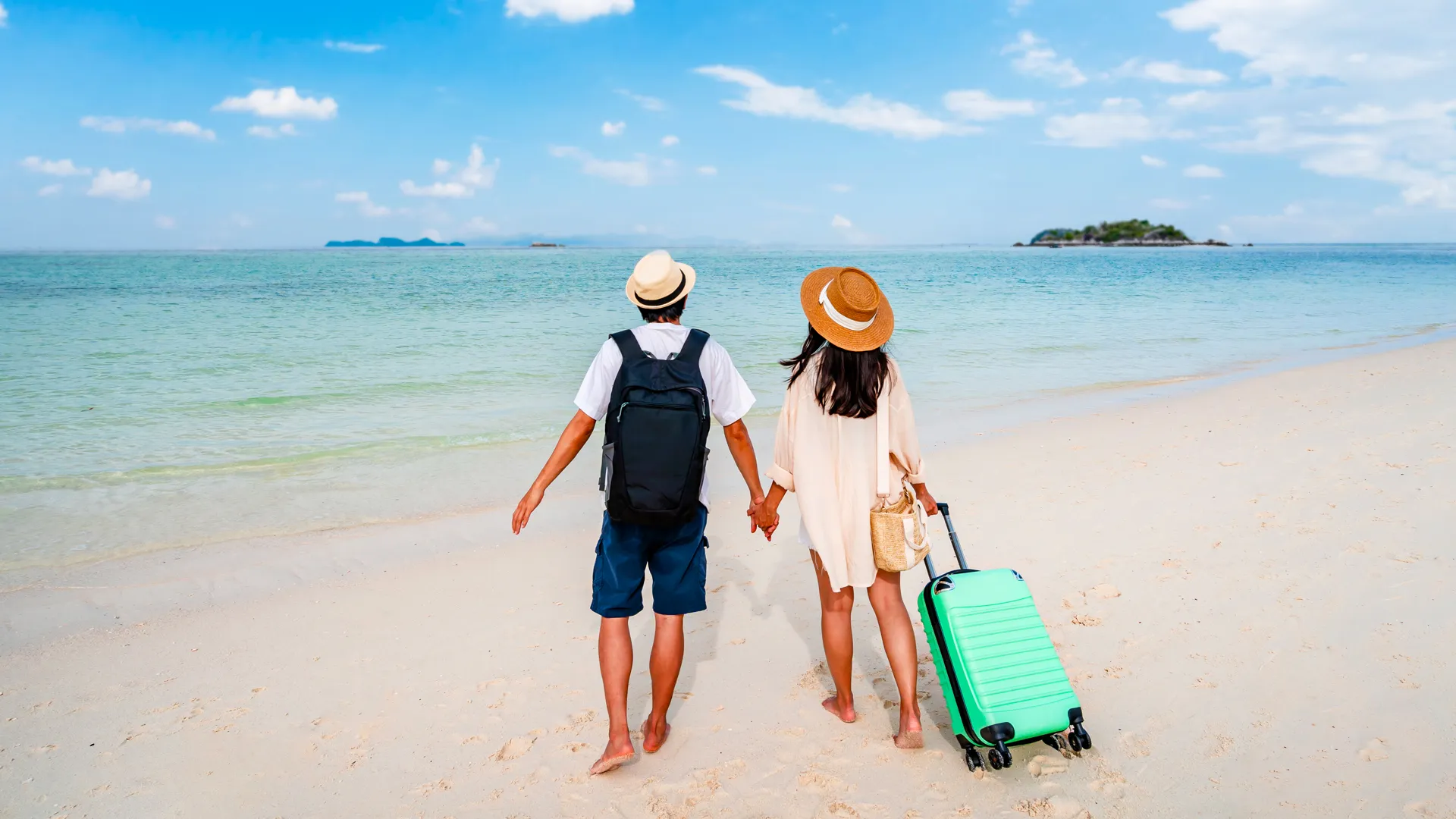 A couple walking hand-in-hand on a white sandy beach at sunset, the man carrying a backpack and wearing a hat, the woman pulling a green suitcase, with turquoise sea and distant islands in the background.