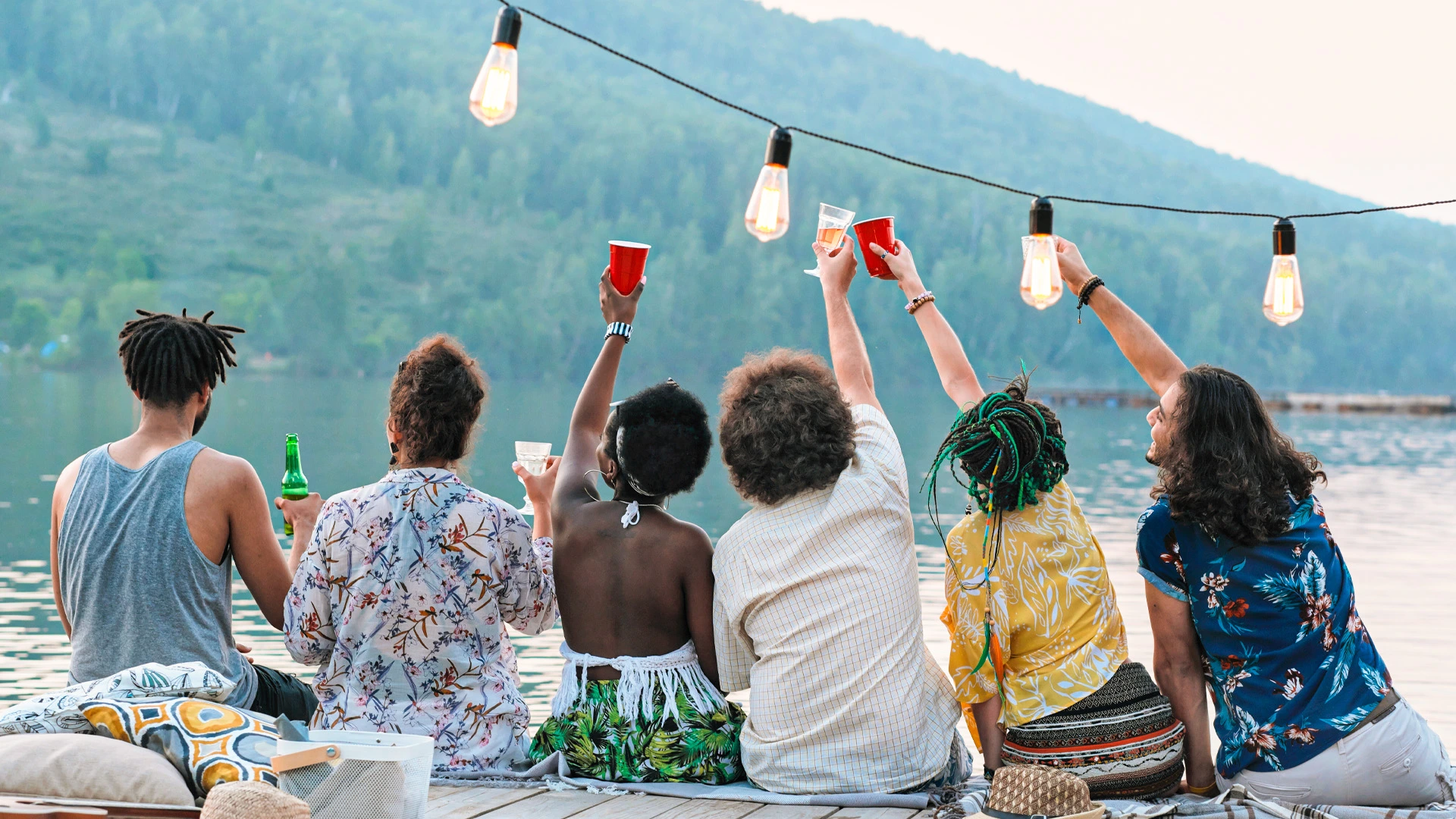 Group of diverse friends sitting on a lakeside deck under string lights, raising drinks together at sunset.