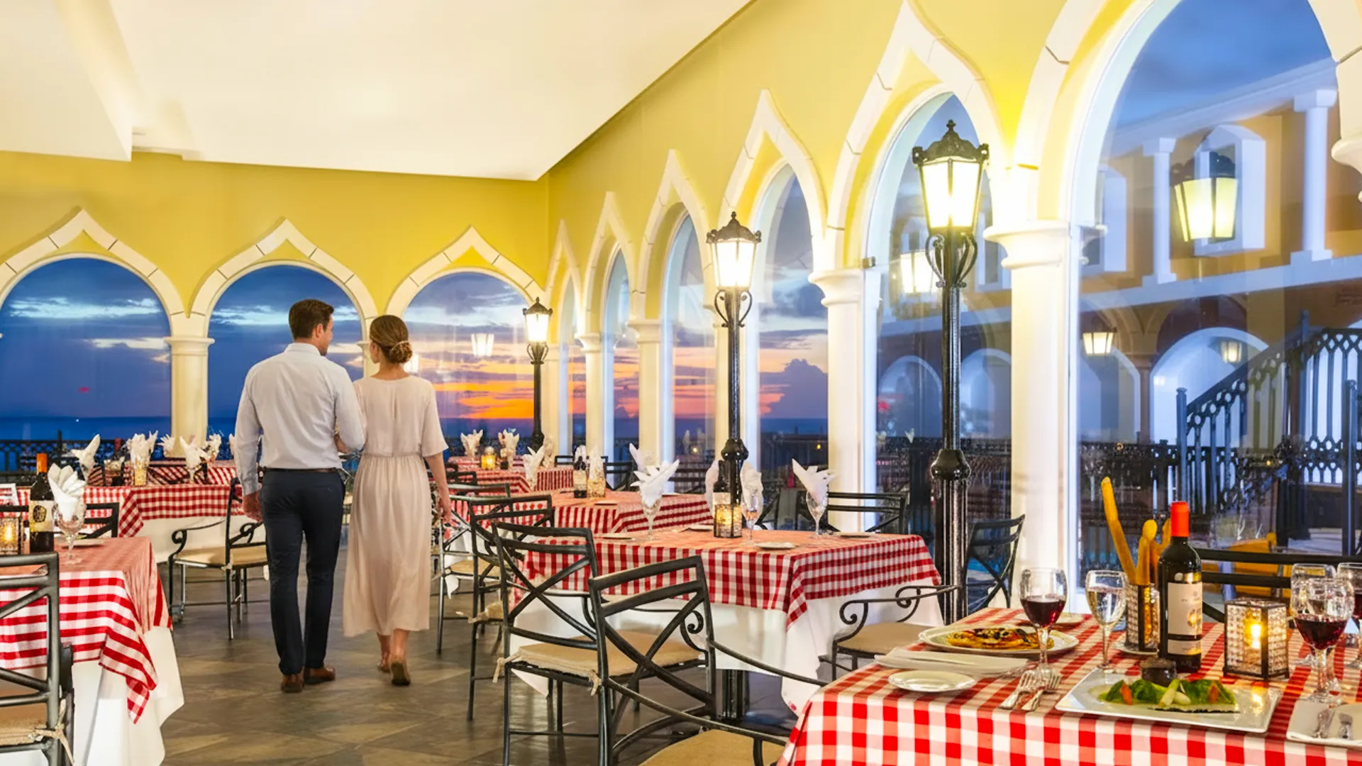 A family of three—father in a dark suit, mother in a light dress, and young child in pink—walking through an elegant indoor restaurant at Crown Paradise Club Cancun, with arched yellow ceilings, red tablecloths, and a sunset view through large windows.