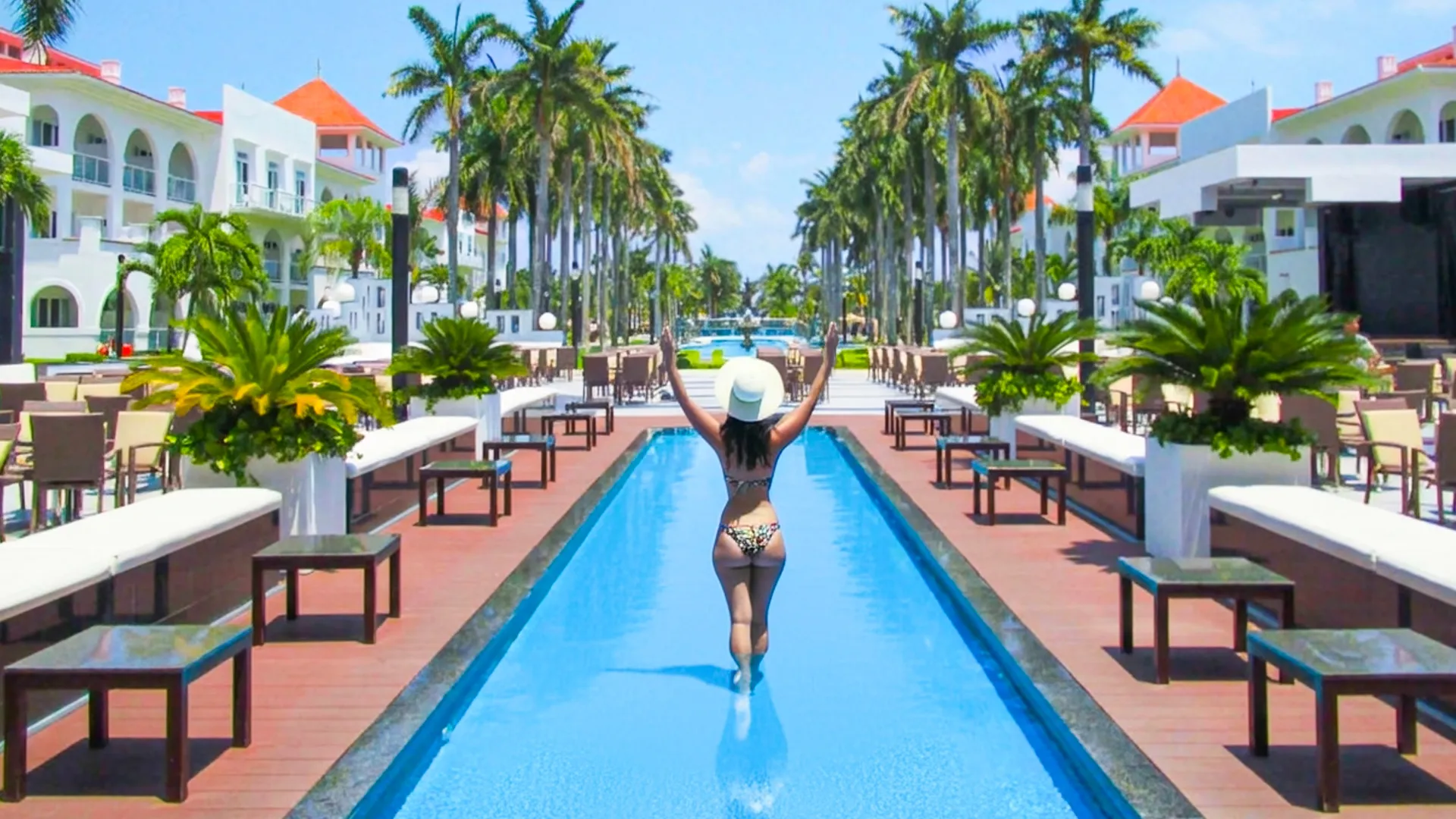 Woman in a hat walking along the central pool lined with palm trees at Riu Palace Cancun.