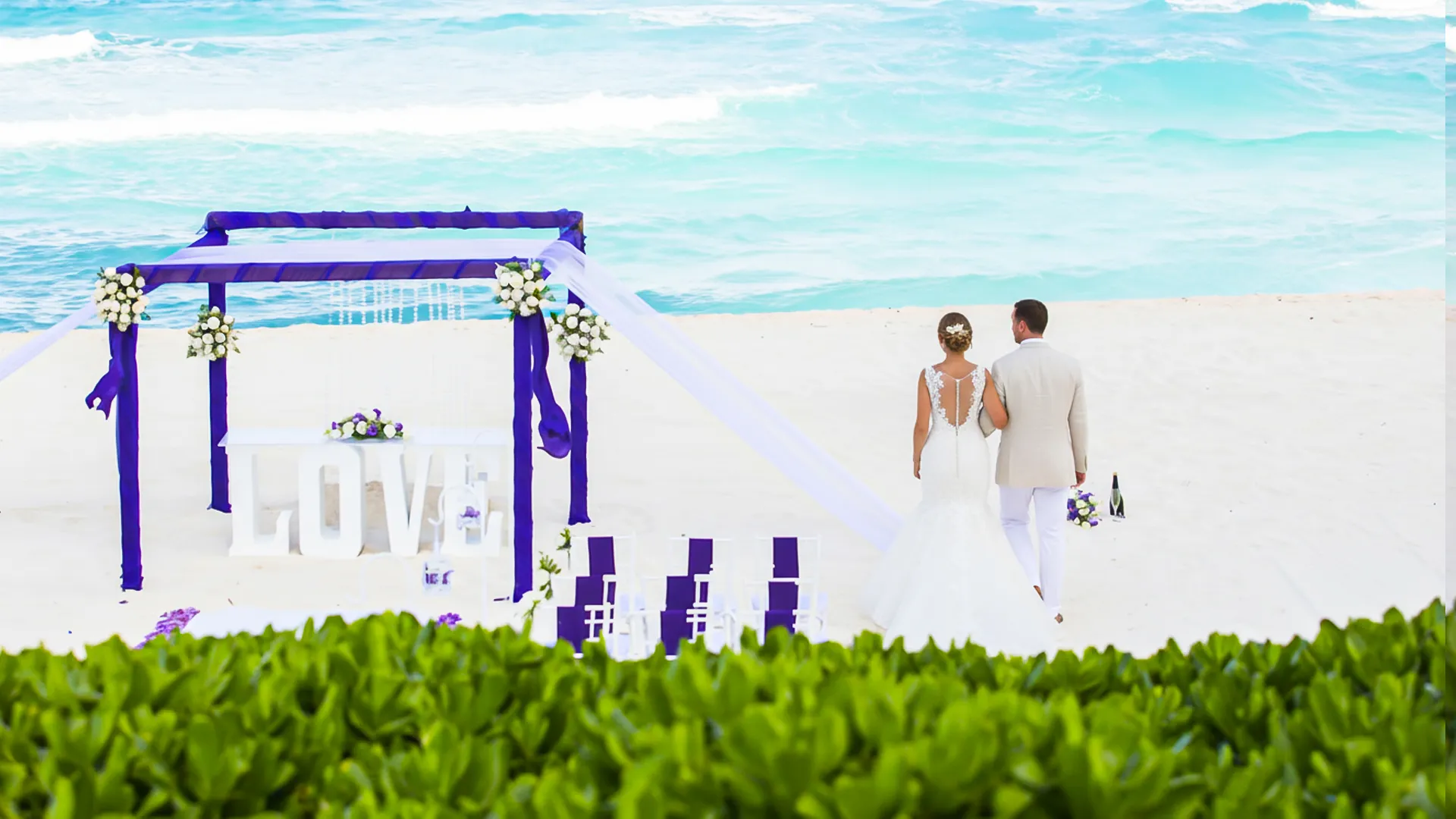 A bride and groom in white attire walking hand-in-hand along a beach at Crown Paradise Club Cancun, with a purple floral arch and white chairs set up for a wedding ceremony, lush greenery, and the turquoise ocean in the background under a clear sky.