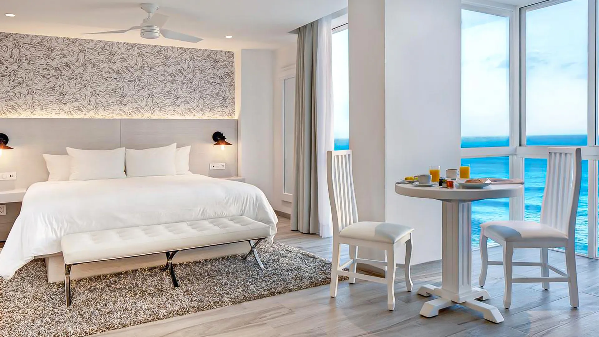 Modern ocean-view suite at Oleo Cancun Playa featuring a king bed, breakfast table, and panoramic windows.