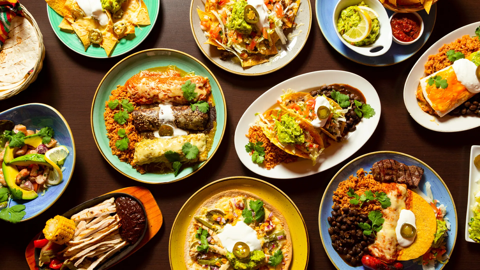 A variety of colorful Mexican dishes including tacos, enchiladas, nachos, and rice served on plates with garnishes like guacamole, salsa, and lime wedges, arranged on a wooden table.