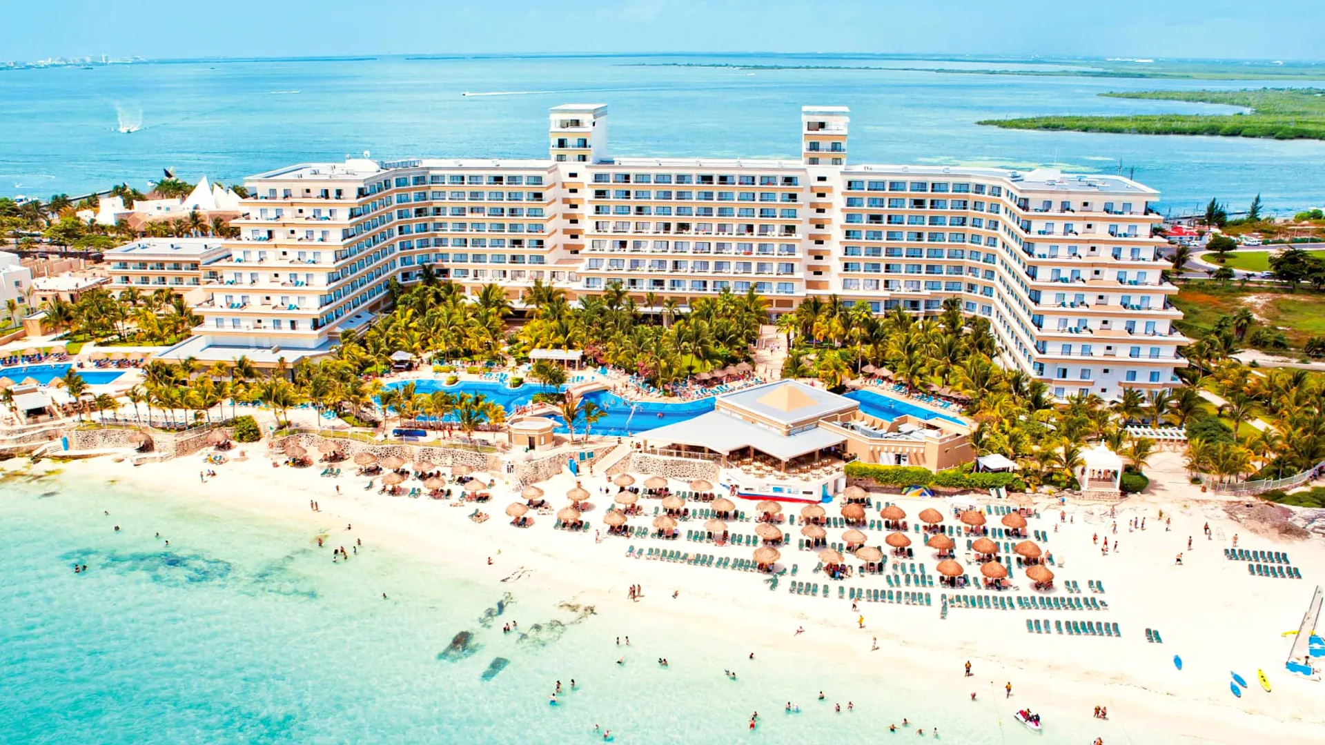 Aerial view of Riu Caribe Cancun showing its beachfront pools, palm gardens, and turquoise shoreline.