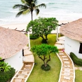 An aerial view of two white thatched-roof bungalows with stone pathways, surrounded by tropical plants and trees, near a sandy beach and ocean waves.