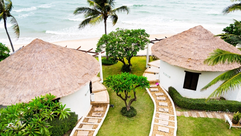 An aerial view of two white thatched-roof bungalows with stone pathways, surrounded by tropical plants and trees, near a sandy beach and ocean waves.