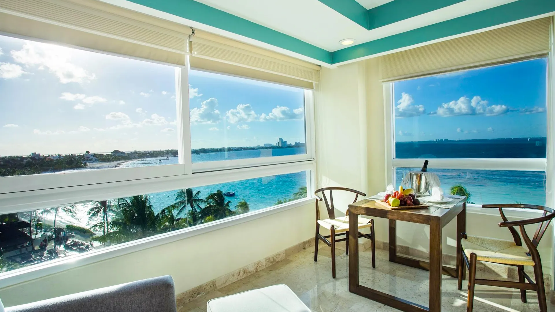 A bright dining area in a Cancun resort suite with floor-to-ceiling windows offering sweeping ocean views, a wooden table set with tropical fruit platters, champagne bottle in an ice bucket, two rattan chairs, beige curtains partially drawn, and fluffy white ottomans on tiled floors under a sunny sky.