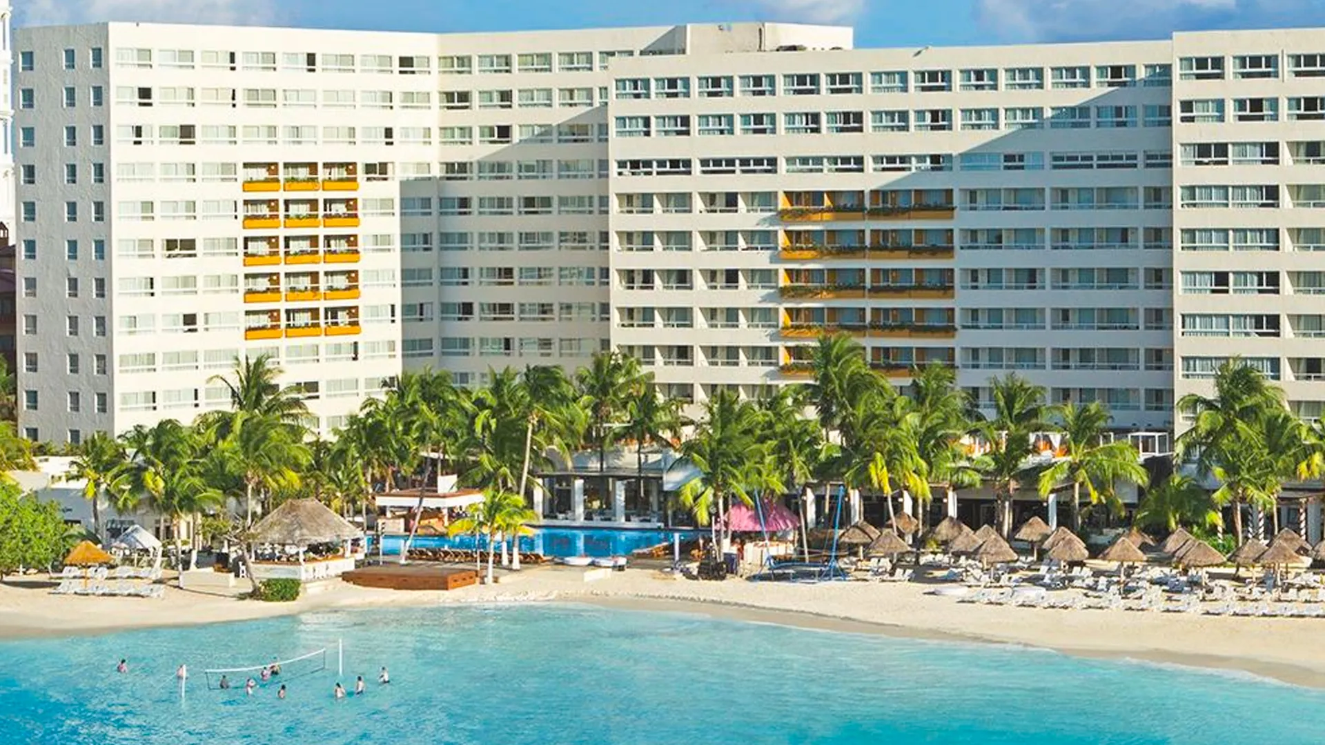 An aerial view of a large Cancun beachfront resort, featuring multiple white high-rise buildings with yellow accents, several outdoor pools including one with volleyball players, lounge chairs on white sand, thatched cabanas, pink umbrellas, tall palm trees, and turquoise ocean waves under a clear blue sky.