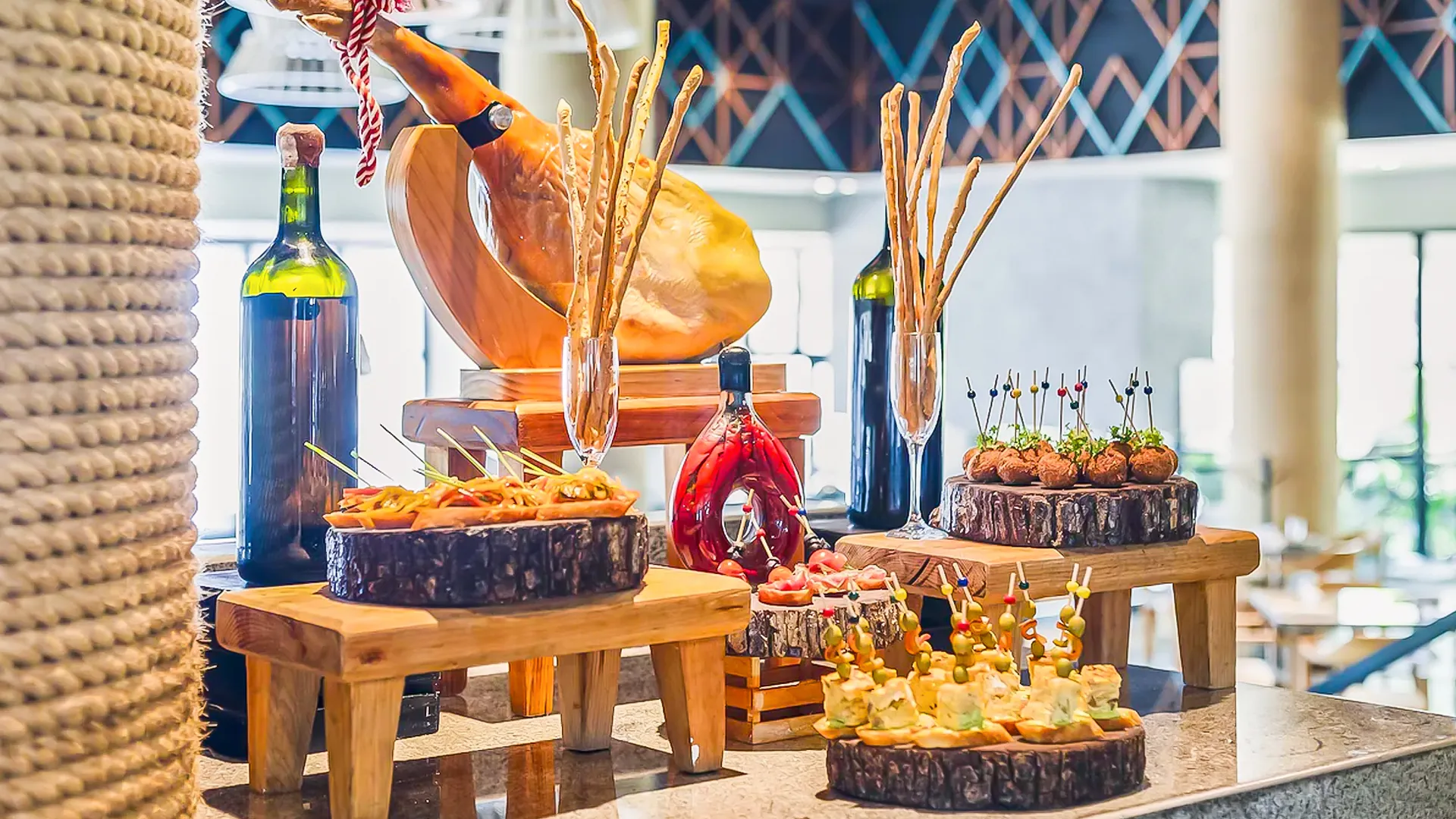 An elegant food display at a Cancun resort buffet, featuring wooden platters with sliced jamón ibérico ham, breadsticks, olive tapenade in small bowls, stuffed mushrooms on skewers, cheese platters, red wine bottles, and decorative elements like striped ribbons and geometric patterns on a marble countertop.