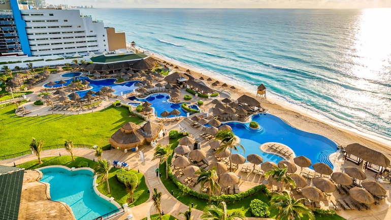 Aerial view of JW Marriott Cancun Resort & Spa at sunset, featuring a modern white high-rise building with blue accents perched on a white sandy beach, multiple interconnected blue pools with thatched-roof palapas and lounge chairs, lush green lawns and palm trees, and the turquoise Caribbean Sea with gentle waves under a golden sky with scattered clouds.