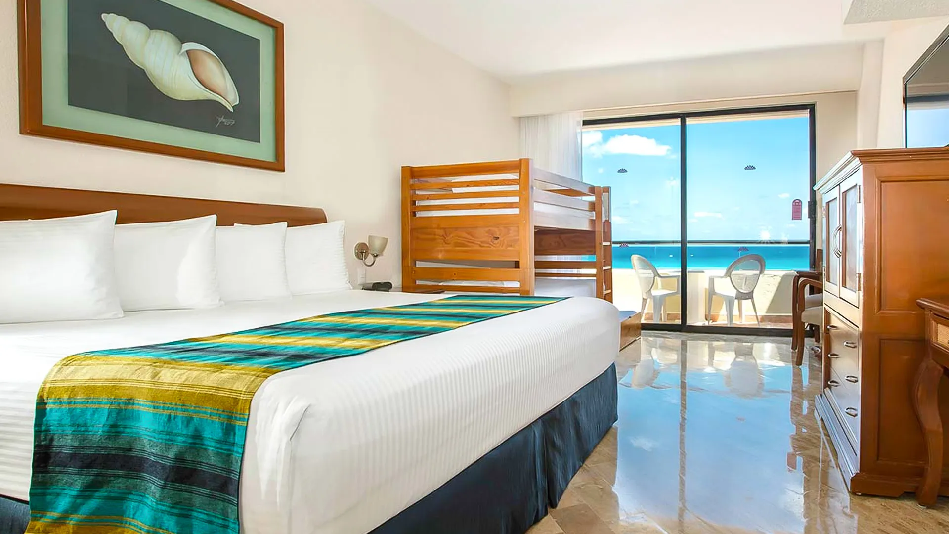 Spacious family suite bedroom at Crown Paradise Club Cancun, showcasing a queen bed with colorful teal and gold striped bedding, a wooden bunk bed with safety rails, white pillows, a framed seashell artwork on the wall, wooden furniture, and floor-to-ceiling sliding doors opening to a balcony with ocean views and lounge chairs on tiled flooring.