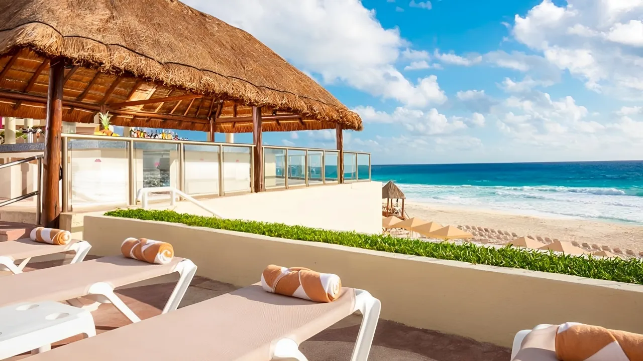 Beachfront swim-up bar at Crown Paradise Club Cancun under a thatched-roof palapa with wooden beams, orange lounge chairs and towels on a tiled deck, potted plants, a bar counter with stools, and a panoramic view of the white sandy beach, turquoise sea, and scattered clouds in the sky.