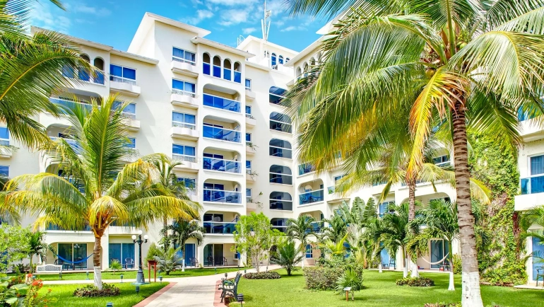 Garden view of Occidental Costa Cancún resort with tropical palm trees, green lawns, and modern white buildings under a bright blue sky.