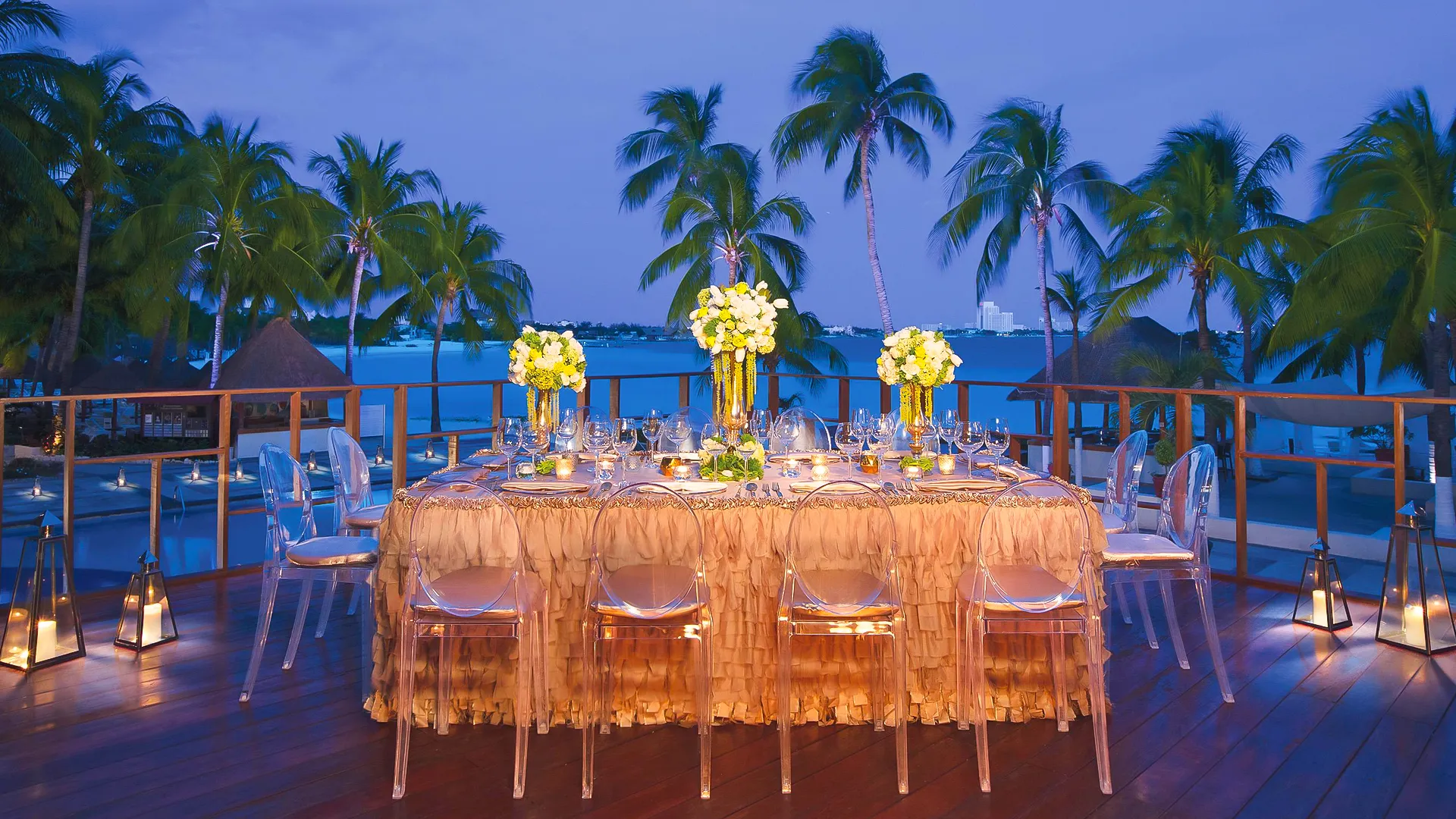An elegant evening dining setup on a wooden terrace at a Cancun resort, featuring a long rectangular table draped in orange fabric with sheer overlays, clear ghost chairs, white floral centerpieces, candlelit lanterns, surrounding palm trees, and a twilight ocean view with distant city lights.