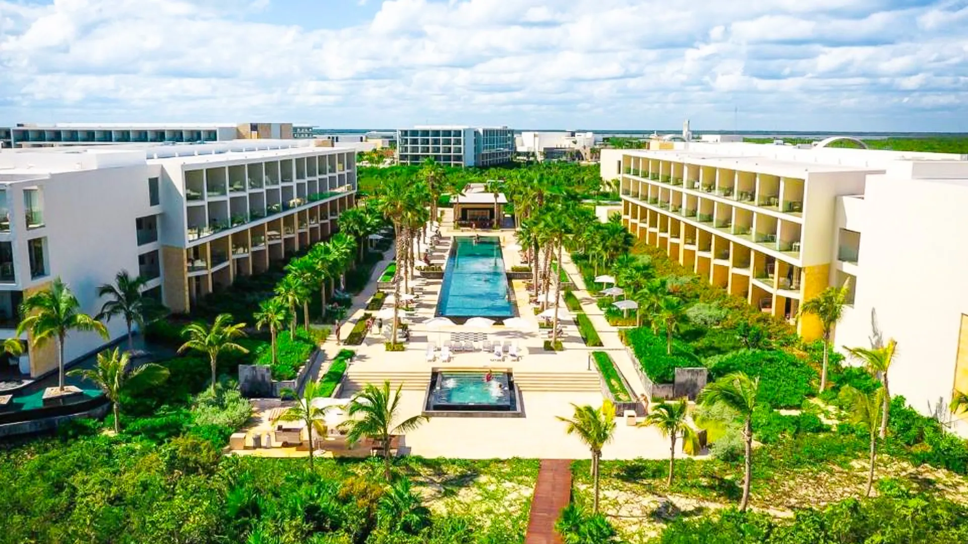 An aerial view of Grand Palladium Cancun Resort showcasing a long infinity pool surrounded by lush tropical gardens, white multi-story buildings, and palm trees under a partly cloudy sky.