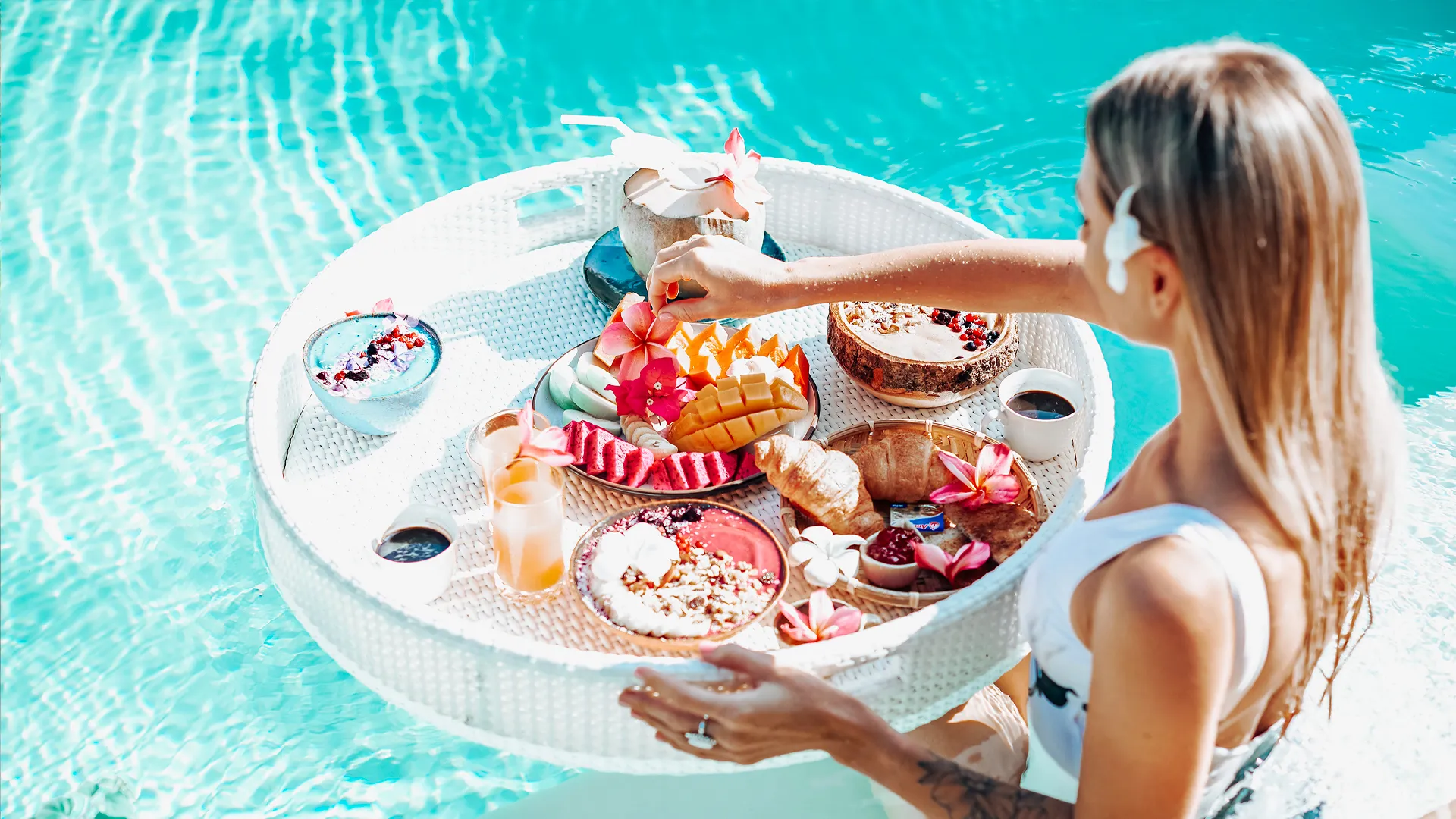 A woman in a white swimsuit floating in a blue pool, serving a large white tray loaded with tropical breakfast items including acai bowls, fresh fruits, croissants, coconut water, and yogurt parfaits.