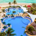 Aerial view of the GR Solaris Cancun resort featuring multiple swimming pools, a multi-story building with red-tiled roofs, palm trees, thatched-roof structures, and a sandy beach with turquoise ocean waters.