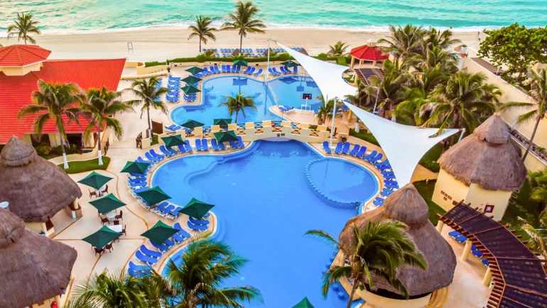 Aerial view of the GR Solaris Cancun resort featuring multiple swimming pools, a multi-story building with red-tiled roofs, palm trees, thatched-roof structures, and a sandy beach with turquoise ocean waters.