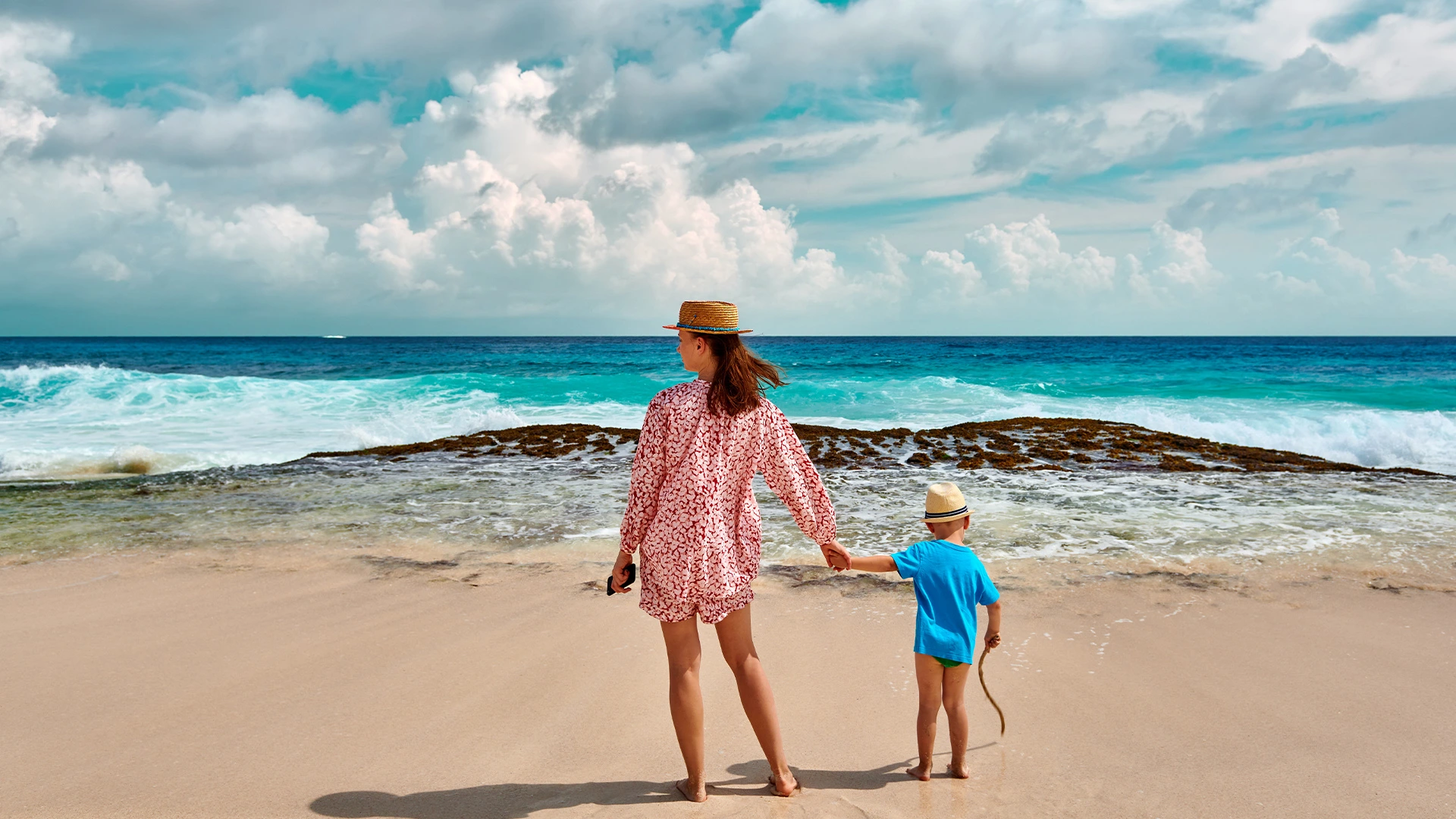 Mother and child standing on a sandy beach looking at turquoise ocean waves under a cloudy blue sky.