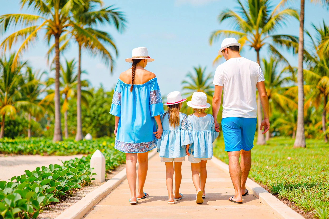 Family of four walking hand-in-hand along a palm-lined path in a tropical resort, dressed in blue and white