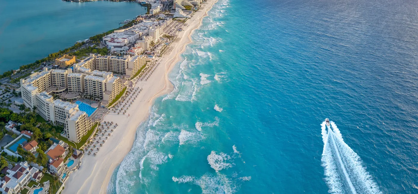 Aerial view of beachfront resorts in Cancun with turquoise water and a speedboat cruising offshore.