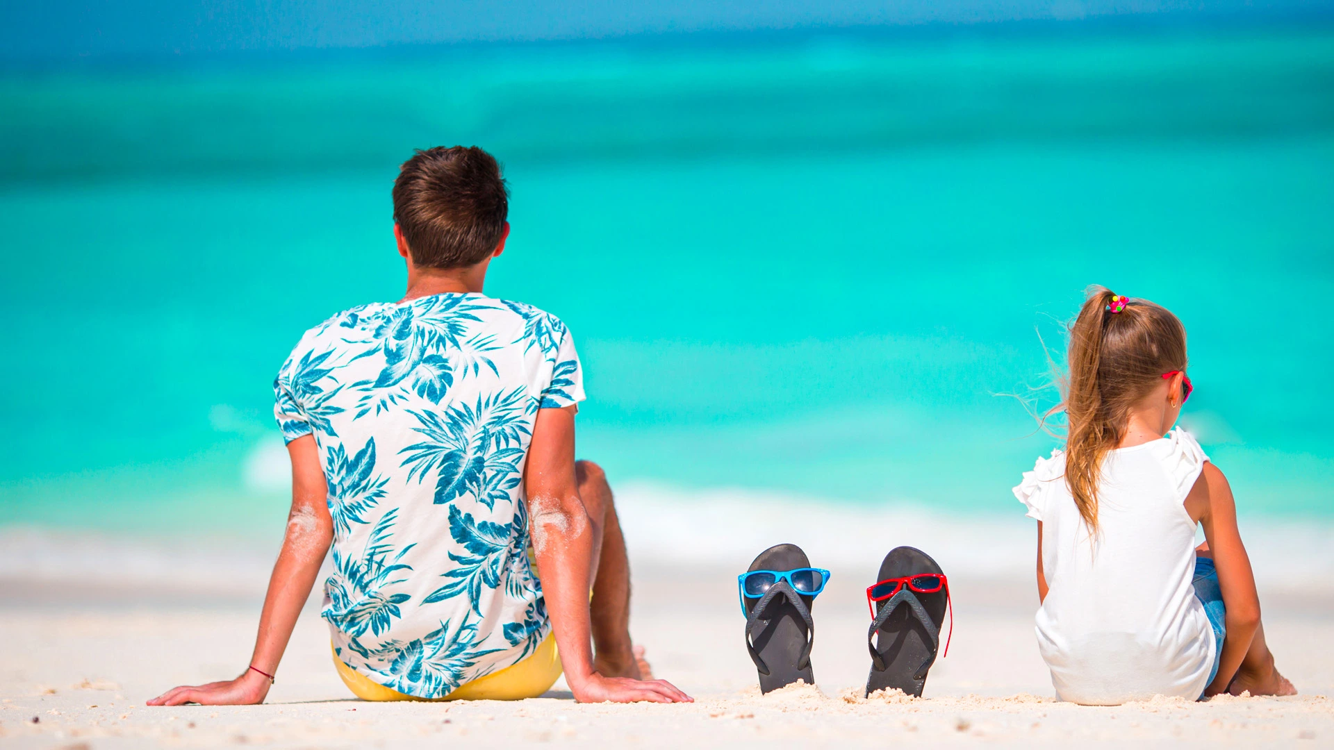 Father and daughter sitting on a white sandy beach facing the turquoise sea with flip-flops beside them.