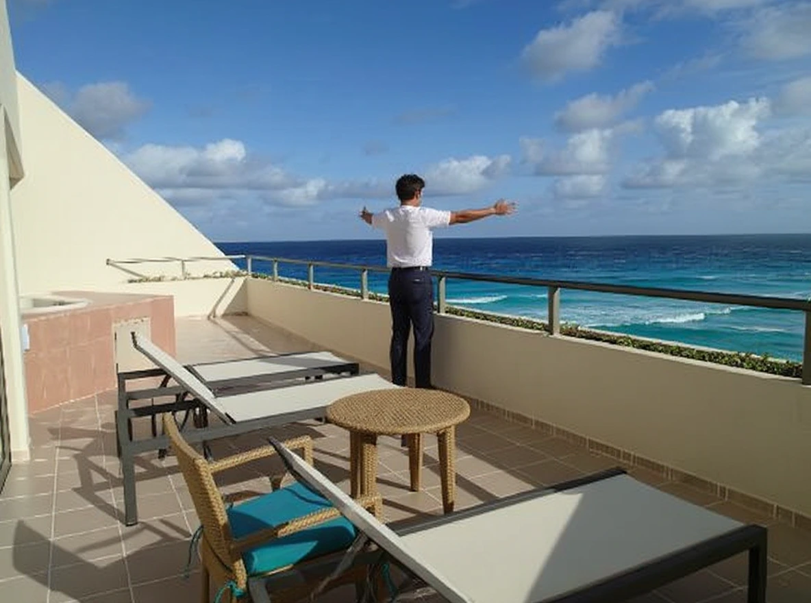 A person standing on a balcony overlooking the ocean, with lounge chairs, a small table, and clear blue skies in the background.
