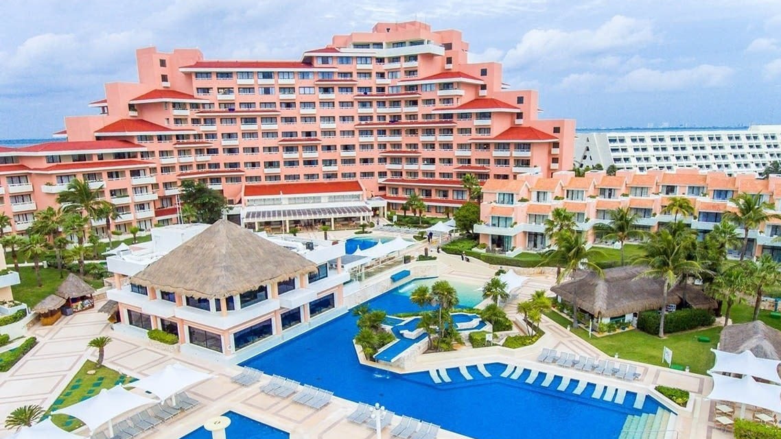Overview of the Wyndham Cancun resort's pink-hued buildings with thatched-roof structures, a rectangular blue swimming pool lined with white lounge chairs, palm trees, tropical landscaping, and partly cloudy skies.