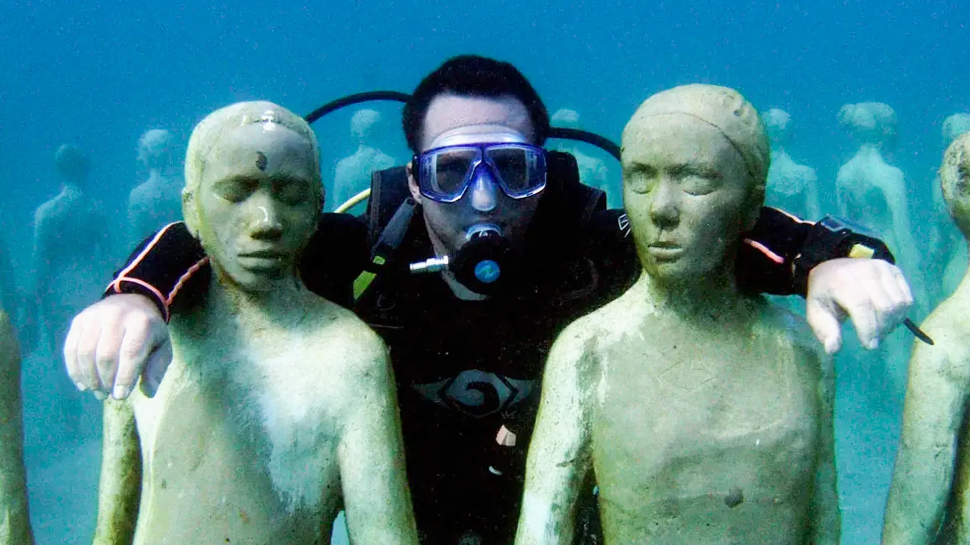 A scuba diver posing with arms around two underwater statues of human figures, set against a clear blue ocean background.