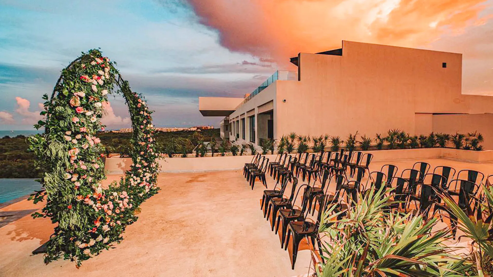 A romantic wedding setup at Atelier Hotel with an arch made of flowers, overlooking the ocean during a beautiful sunset.
