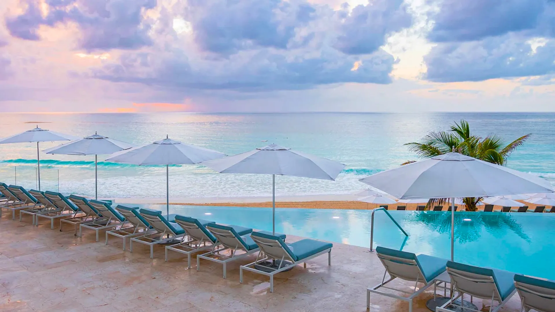 Sun Palace Cancun poolside with lounge chairs and umbrellas facing the beach, with a view of the beautiful turquoise ocean and the sky filled with pastel colors at sunset.